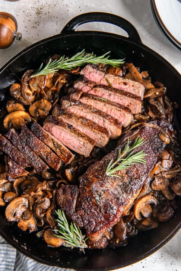 New York strip steaks with sautéed mushrooms and onions in a cast iron skillet, with one steak sliced to show a medium-rare center and garnished with fresh rosemary.