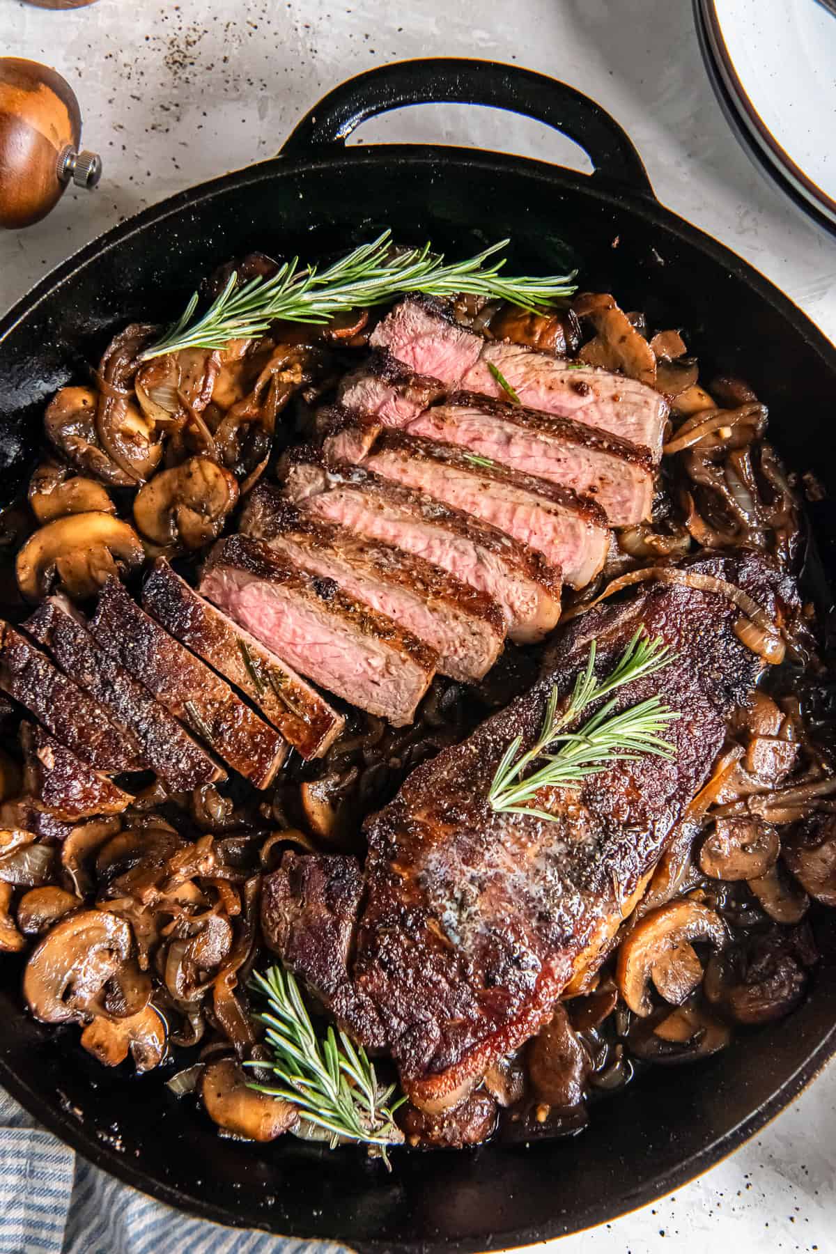 New York strip steaks with sautéed mushrooms and onions in a cast iron skillet, with one steak sliced to show a medium-rare center and garnished with fresh rosemary.