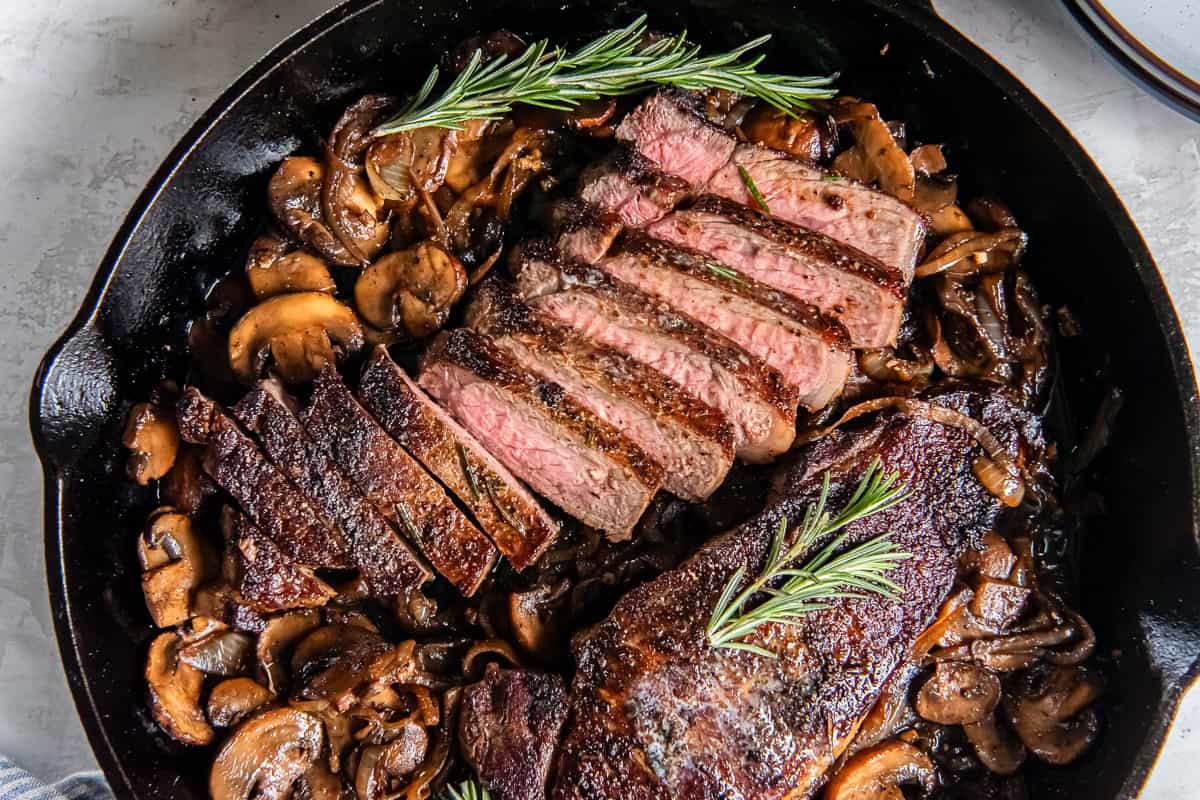 New York strip steaks with sautéed mushrooms and onions in a cast iron skillet, with one steak sliced to show a juicy medium-rare center and fresh rosemary garnish.