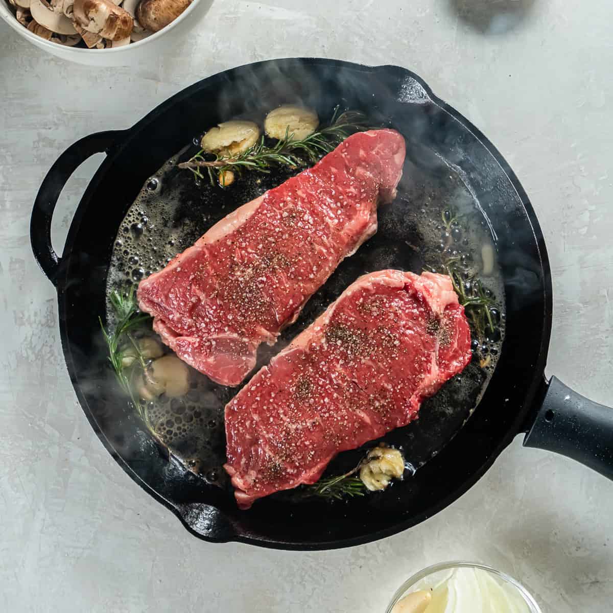 New York strip steaks searing in a cast iron skillet with rosemary, garlic, and butter.