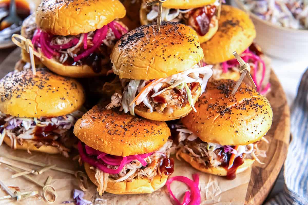 Pulled pork sliders topped with creamy coleslaw, barbecue sauce, and pickled red onions on a serving board.