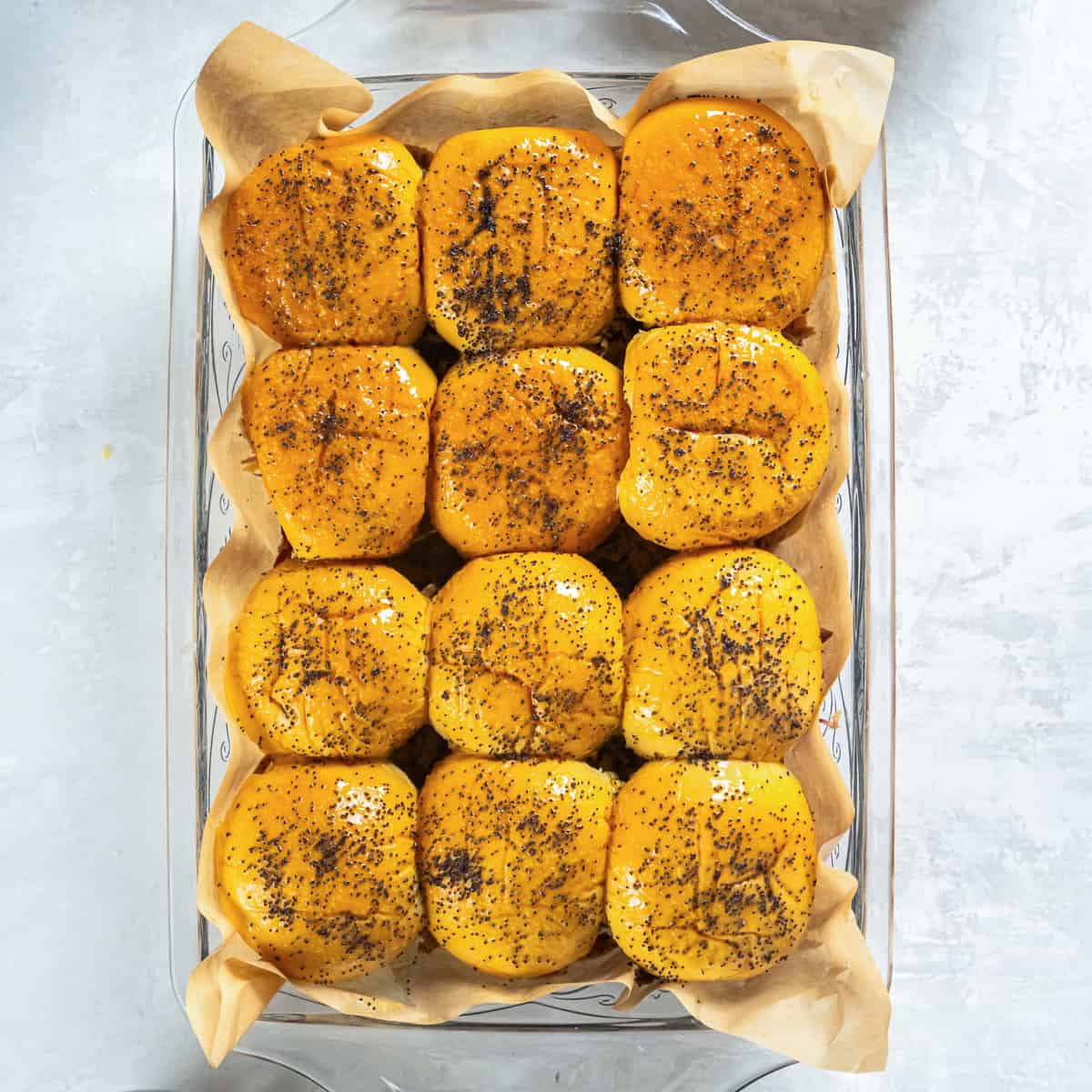 Pulled pork sliders baked in a baking dish with butter-brushed slider rolls topped with poppy seeds.