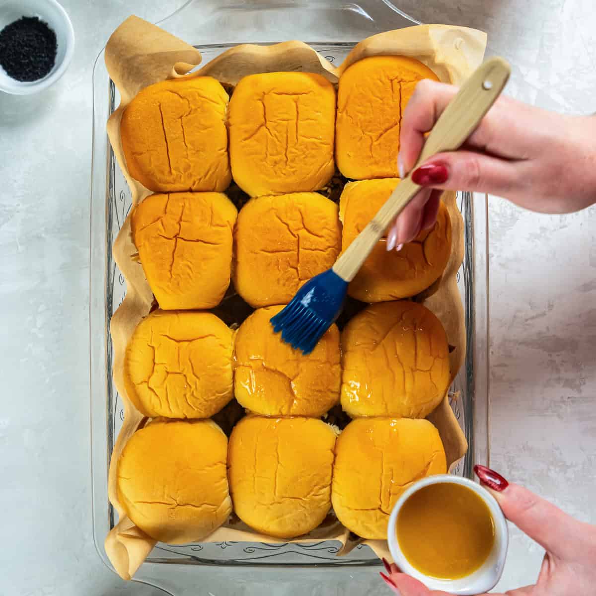 Brushing melted butter over slider rolls in a baking dish before sprinkling with poppy seeds and baking.