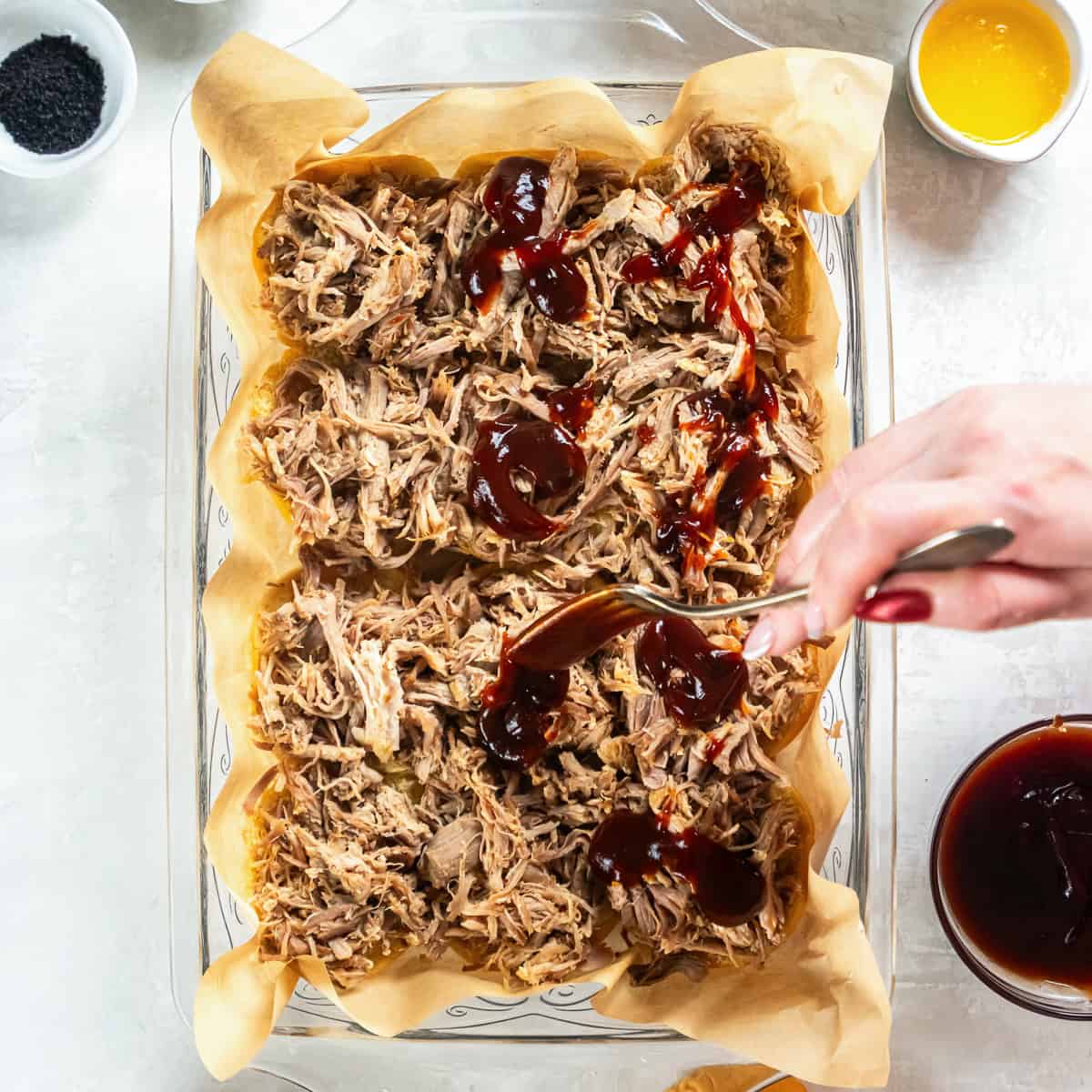 Barbecue sauce being spooned over shredded pulled pork layered on slider rolls in a baking dish.