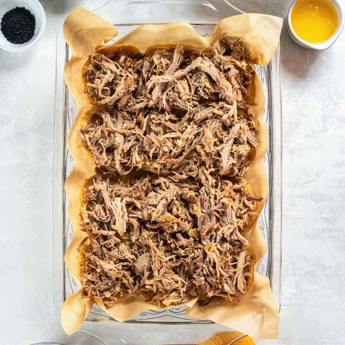 Shredded pulled pork layered over slider rolls in a parchment-lined baking dish for pulled pork sliders.