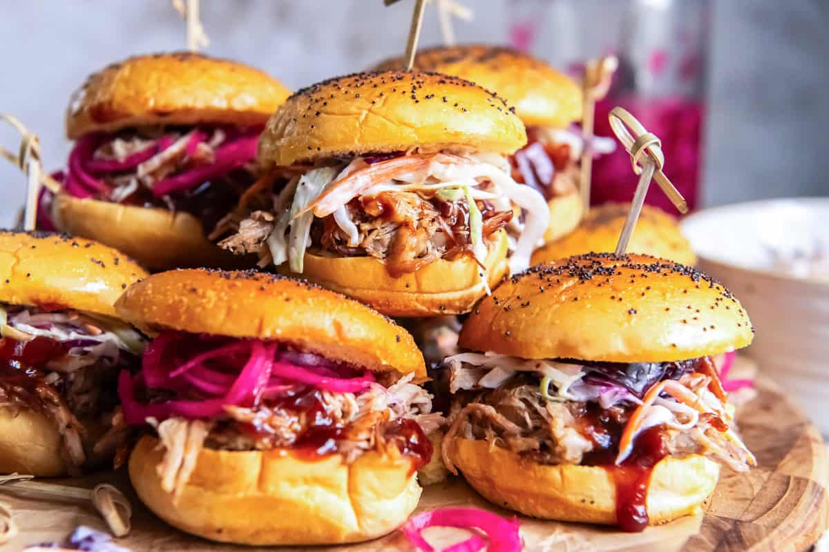 Pulled pork sliders topped with creamy coleslaw, barbecue sauce, and pickled red onions on a serving board.