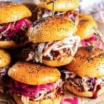 Pulled pork sliders topped with creamy coleslaw and pickled red onions, arranged on a serving board with barbecue sauce and a jar of pickled onions in the background.
