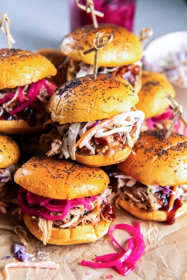 Pulled pork sliders topped with creamy coleslaw and pickled red onions, arranged on a serving board with barbecue sauce and a jar of pickled onions in the background.