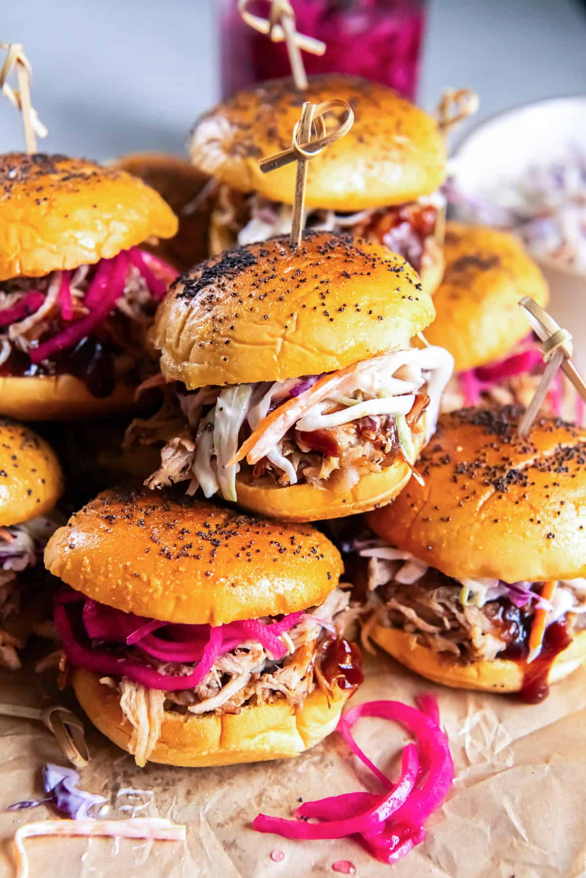 Pulled pork sliders topped with creamy coleslaw and pickled red onions, arranged on a serving board with barbecue sauce and a jar of pickled onions in the background.