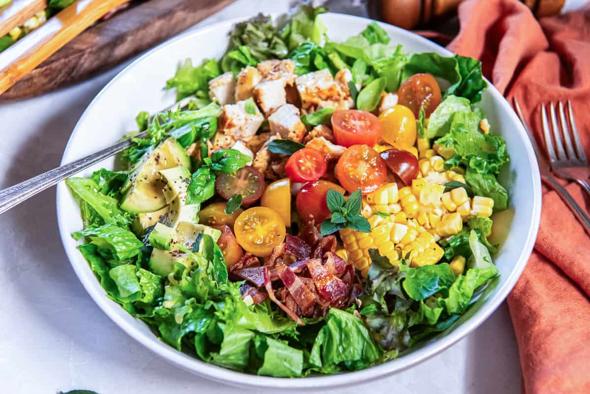 Summer chicken salad with chicken, avocado, bacon, sweet corn, cherry tomatoes, and mixed greens, garnished with fresh herbs and served with vinaigrette dressing.