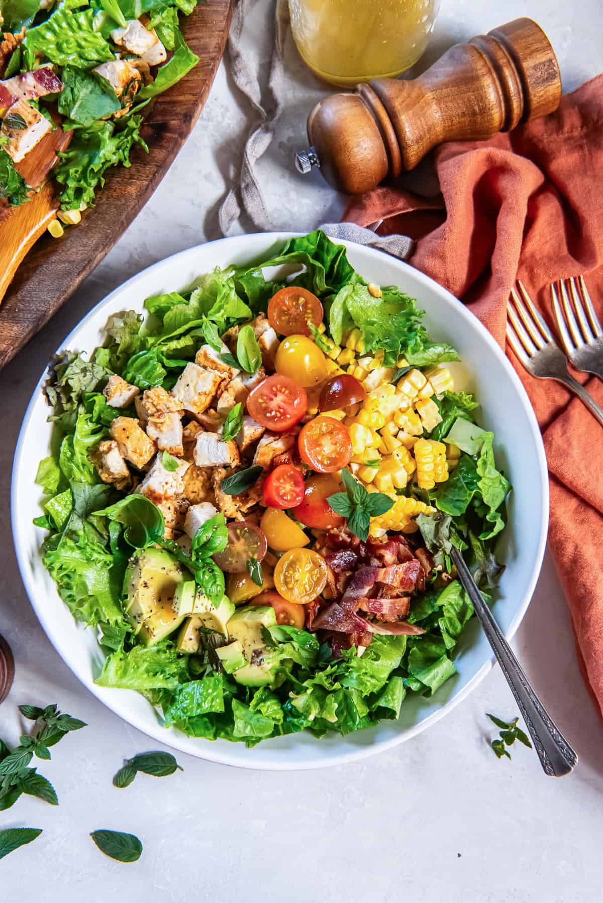 Plated summer chicken salad made with grilled chicken, avocado, sweet corn, bacon, cherry tomatoes, and mixed greens, topped with fresh herbs and served with a homemade vinaigrette.
