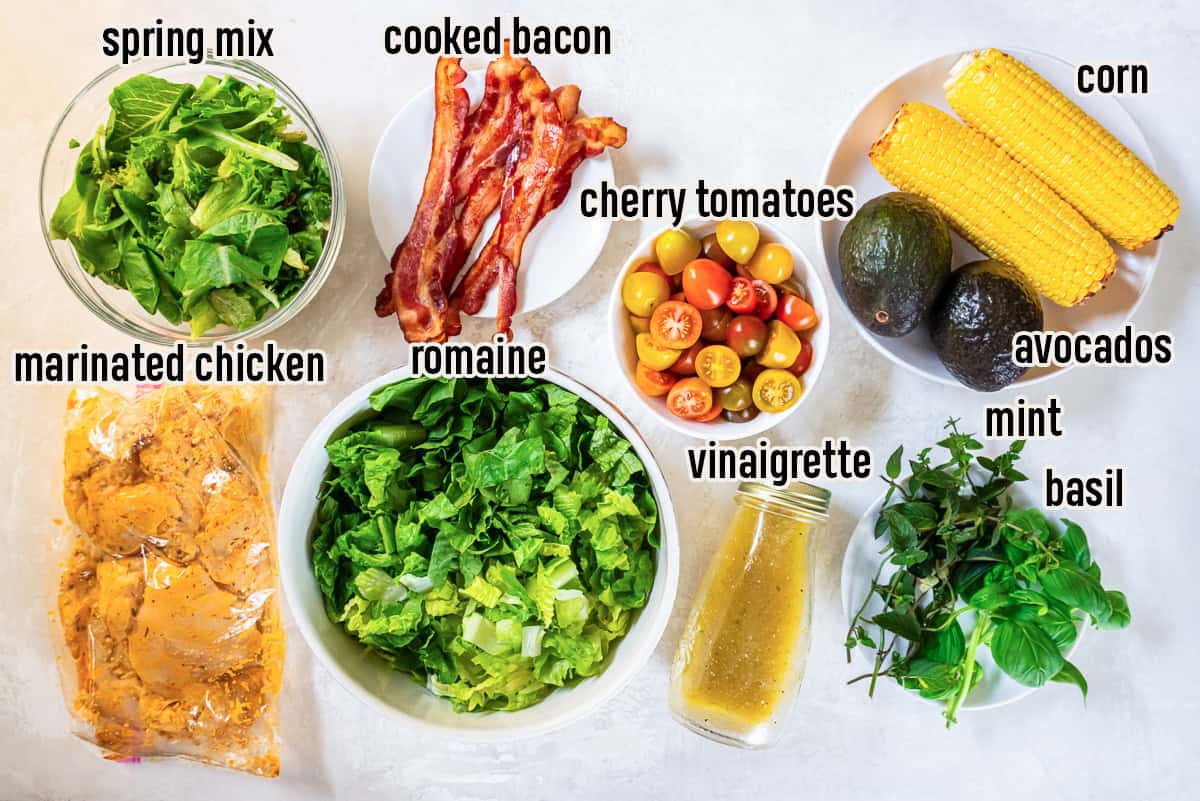 Ingredients for Summer Chicken Salad with Bacon, Corn, and Avocado including marinated chicken, bacon, romaine lettuce, spring mix, cherry tomatoes, fresh corn, avocados, basil, mint, and apple cider vinaigrette.