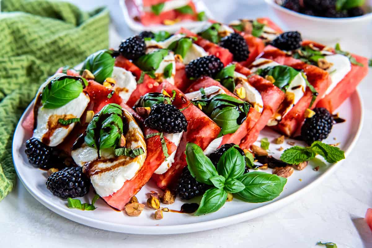 Watermelon Caprese arranged on a white platter with layers of watermelon, fresh mozzarella, basil, blackberries, pistachios, and a drizzle of balsamic glaze.