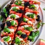 Watermelon Caprese on a serving platter with layers of watermelon, fresh mozzarella, basil leaves, blackberries, chopped pistachios, and balsamic glaze.