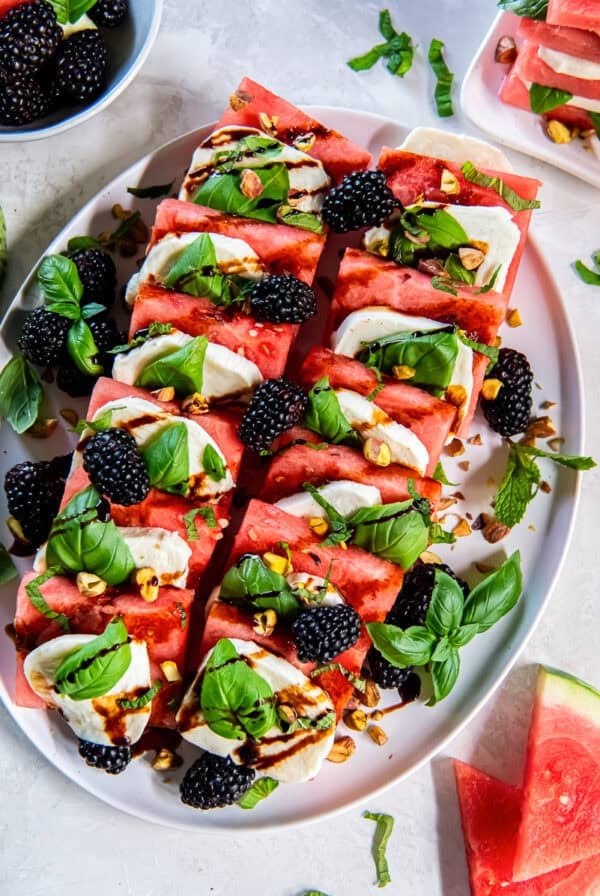 Watermelon Caprese on a serving platter with layers of watermelon, fresh mozzarella, basil leaves, blackberries, chopped pistachios, and balsamic glaze.