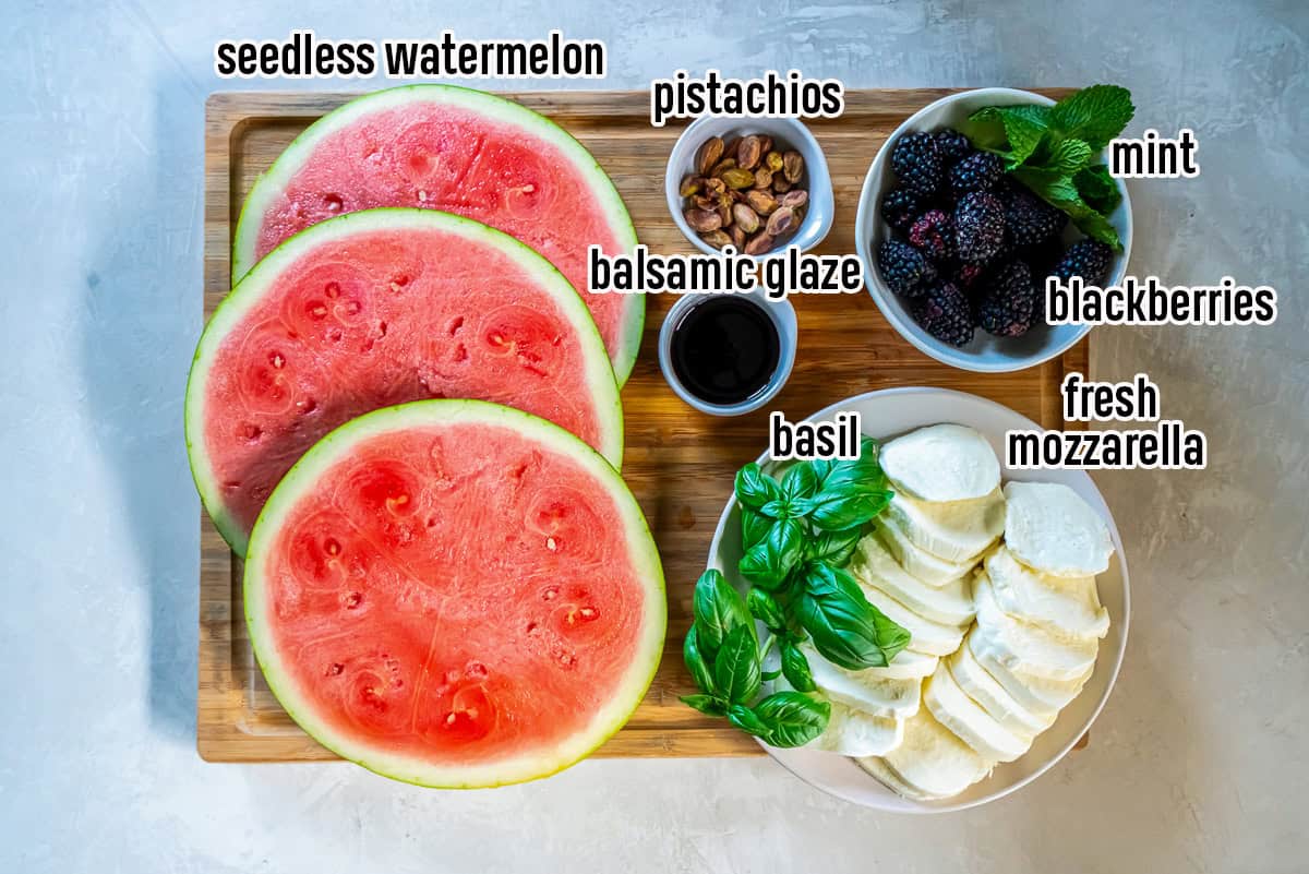 Ingredients for watermelon caprese salad including sliced seedless watermelon, fresh mozzarella, basil, blackberries, pistachios, mint, and balsamic glaze arranged on a wooden board.