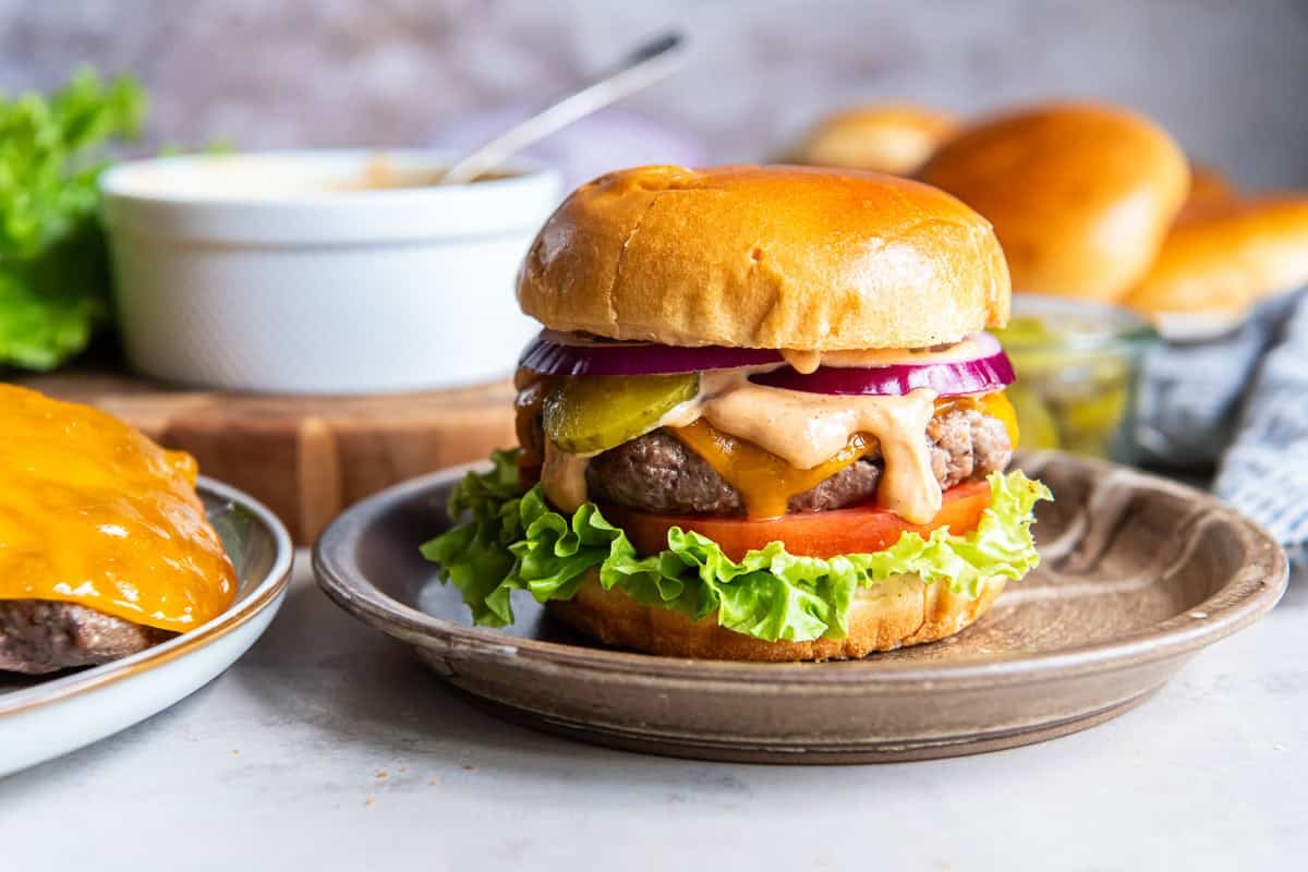 Homemade cheeseburger topped with creamy burger sauce, cheddar cheese, lettuce, tomato, pickles, and red onion on a toasted bun.