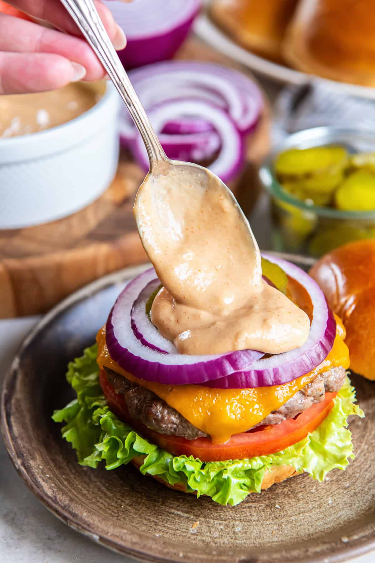 Spoon adding homemade burger sauce to a cheeseburger with lettuce, tomato, pickles, and red onion.