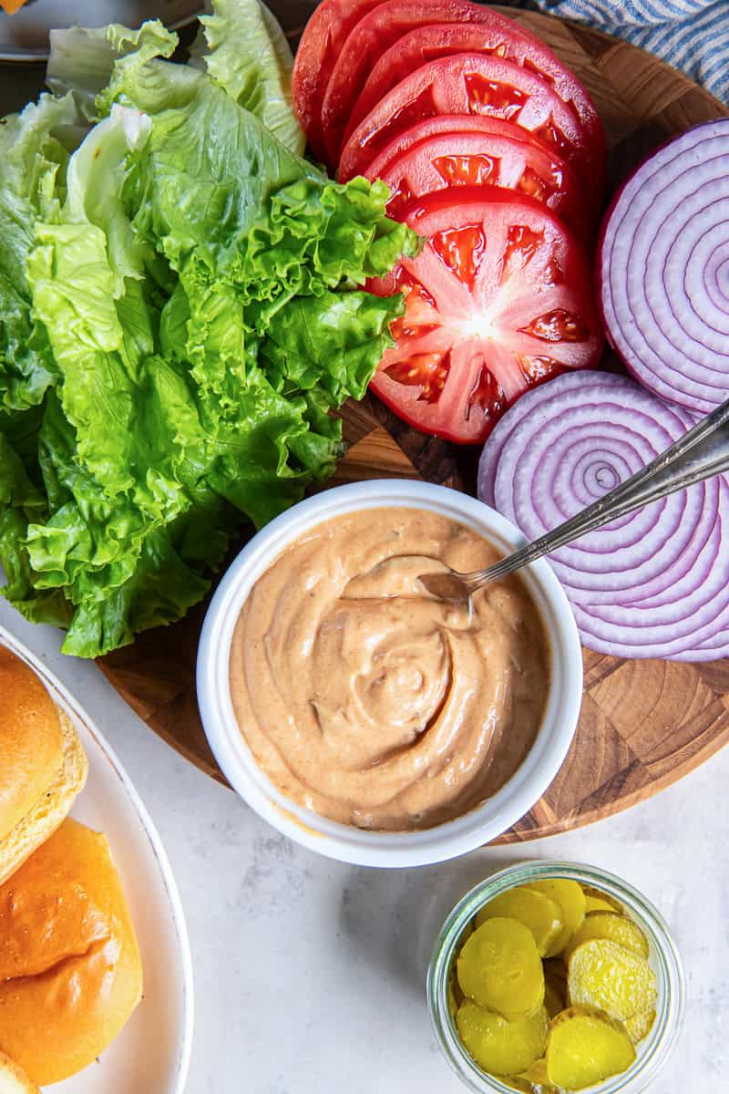 Homemade burger sauce in a white bowl surrounded by lettuce, tomato slices, red onion, pickles, and burger buns.