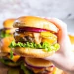 Hand holding a cheeseburger topped with homemade burger sauce, cheddar cheese, pickles, red onion, lettuce, and tomato.
