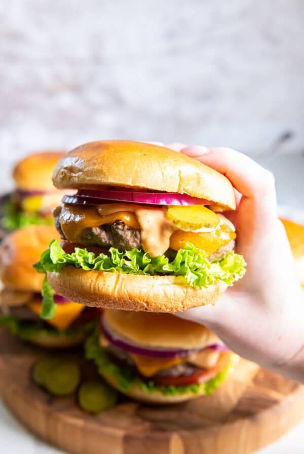 Hand holding a cheeseburger topped with homemade burger sauce, cheddar cheese, pickles, red onion, lettuce, and tomato.