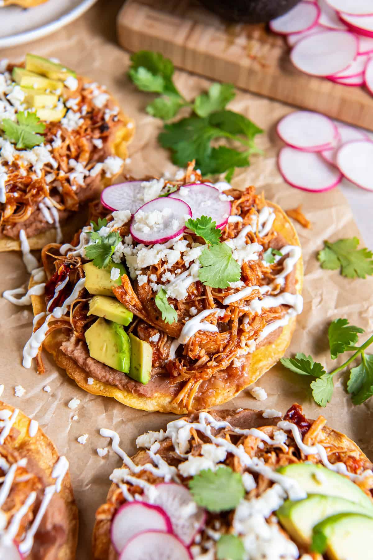 Chicken Tinga tostada topped with refried beans, avocado, sliced radishes, Cotija cheese, cilantro, and Mexican crema, with additional tostadas blurred in the background.