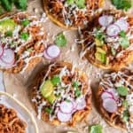 Chicken Tinga tostadas topped with refried beans, avocado, radishes, Cotija cheese, cilantro, and Mexican crema, served with a bowl of Chicken Tinga and fresh lime wedges.