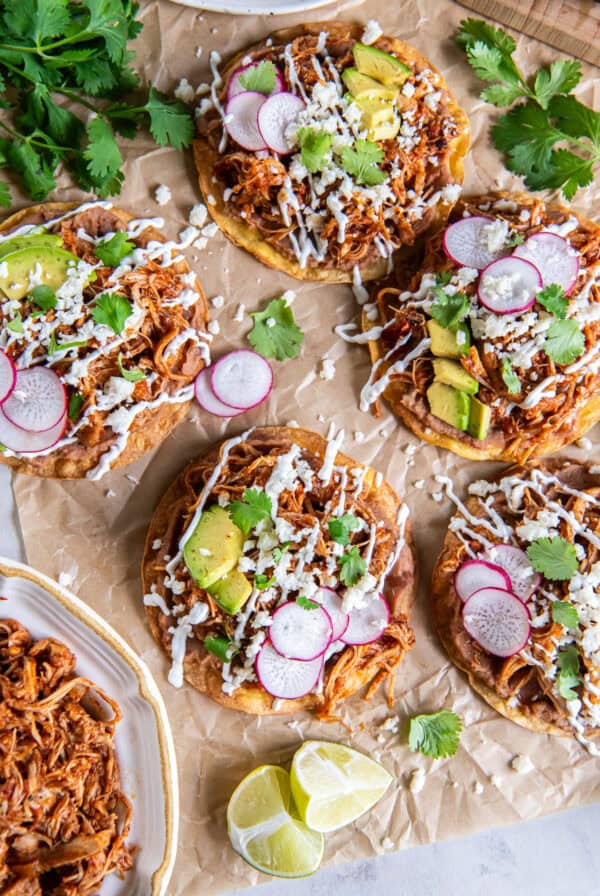 Chicken Tinga tostadas topped with refried beans, avocado, radishes, Cotija cheese, cilantro, and Mexican crema, served with a bowl of Chicken Tinga and fresh lime wedges.