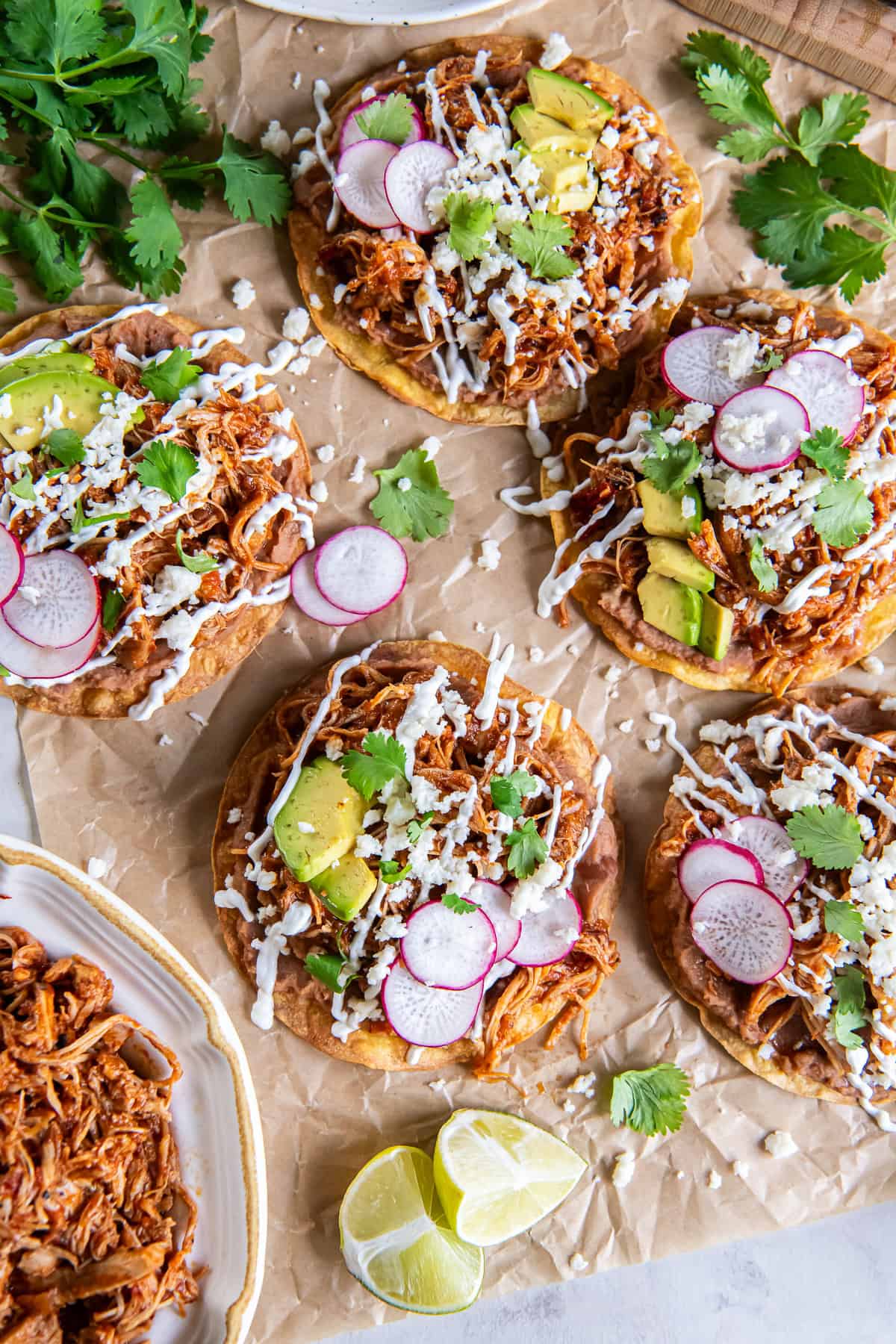Chicken Tinga tostadas topped with refried beans, avocado, radishes, Cotija cheese, cilantro, and Mexican crema, served with a bowl of Chicken Tinga and fresh lime wedges.