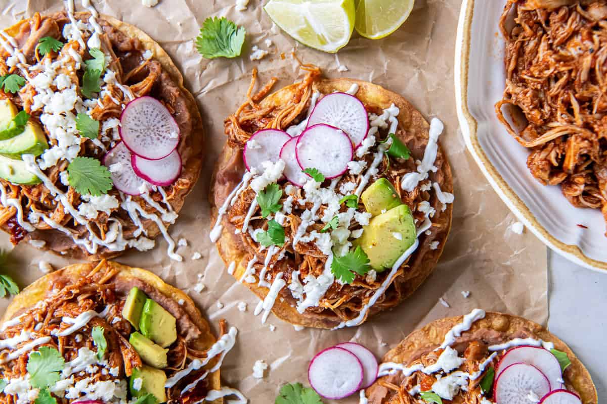 Chicken Tinga tostadas topped with refried beans, avocado, sliced radishes, Cotija cheese, cilantro, and Mexican crema, served with a bowl of Chicken Tinga and fresh lime wedges.