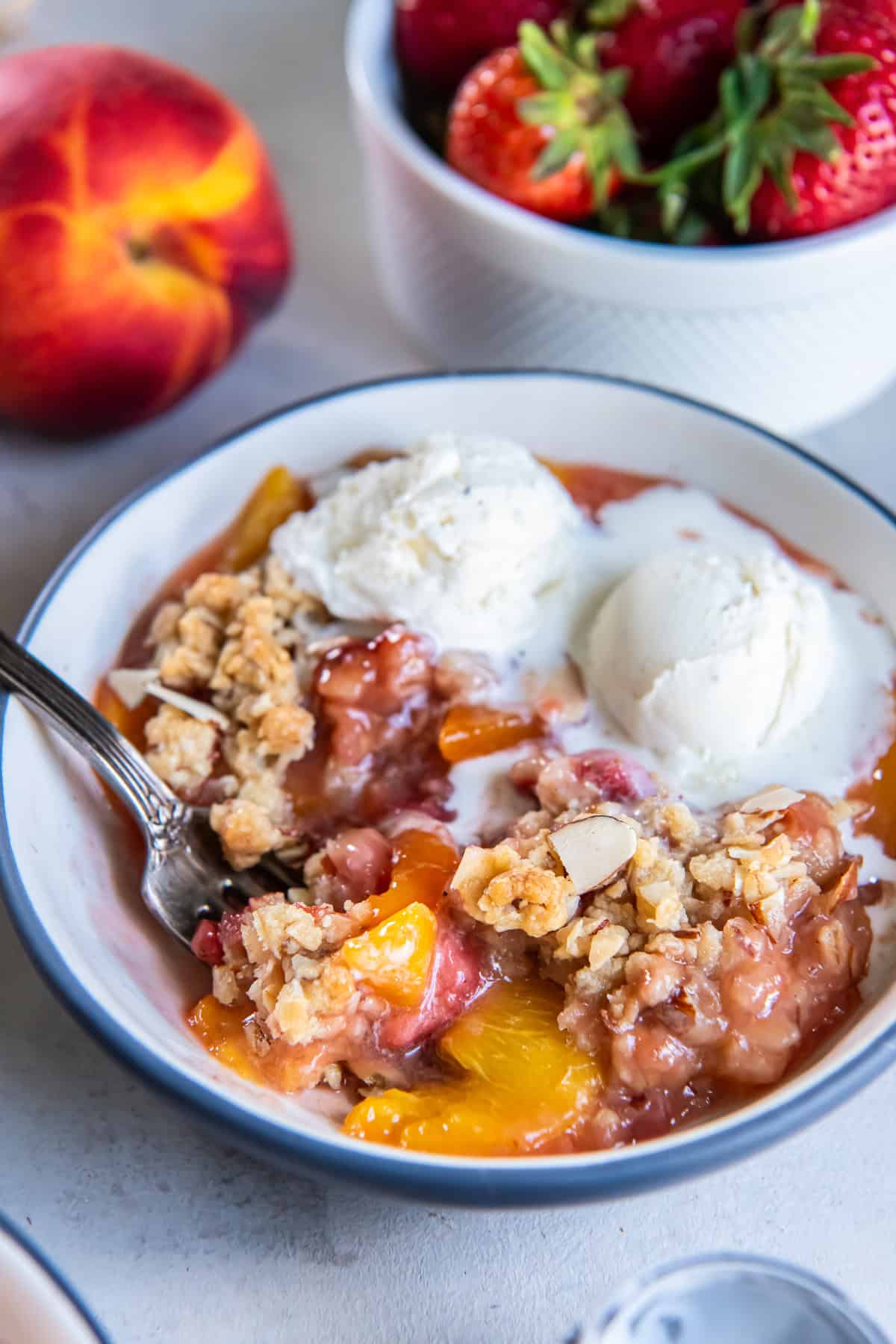 Bowl of warm Peach Strawberry Crisp topped with vanilla ice cream and almond oat crumble, with fresh peaches and strawberries in the background.