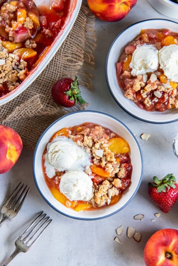 Peach Strawberry Crisp with vanilla ice cream served in bowls next to the baking dish and fresh peaches and strawberries.
