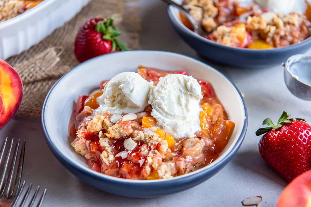 Peach Strawberry Crisp served warm with vanilla ice cream in a bowl, with fresh peaches and strawberries in the background.