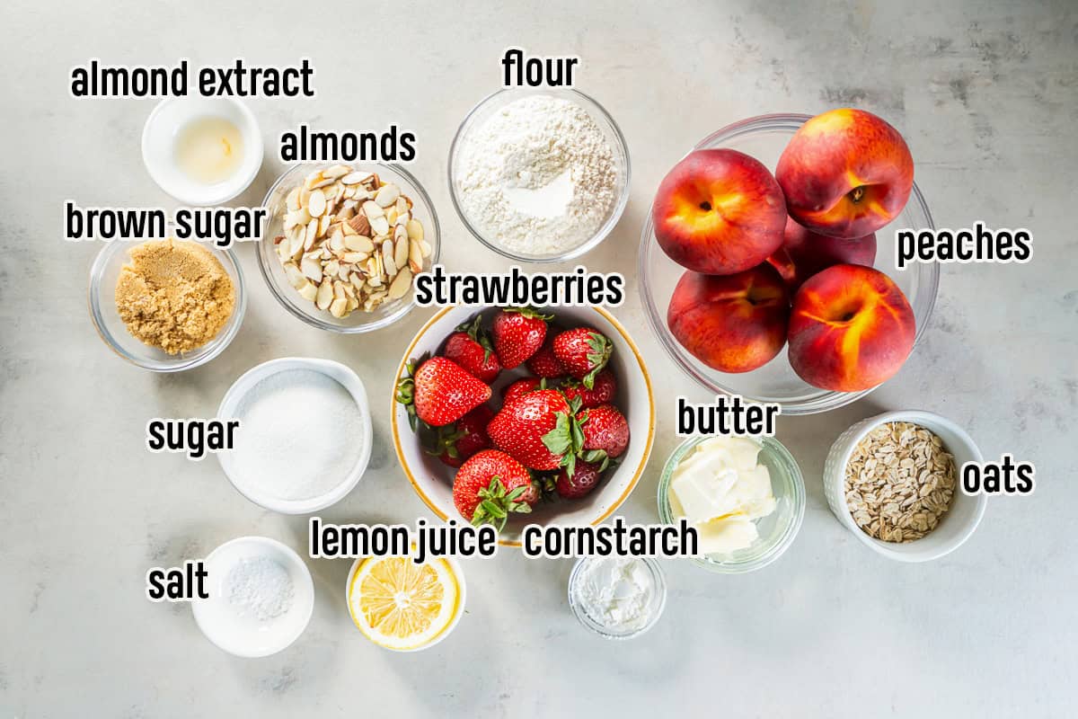 Ingredients for Peach Strawberry Crisp arranged on a countertop, including fresh peaches, strawberries, oats, flour, sliced almonds, butter, sugars, lemon juice, almond extract, cornstarch, and salt.