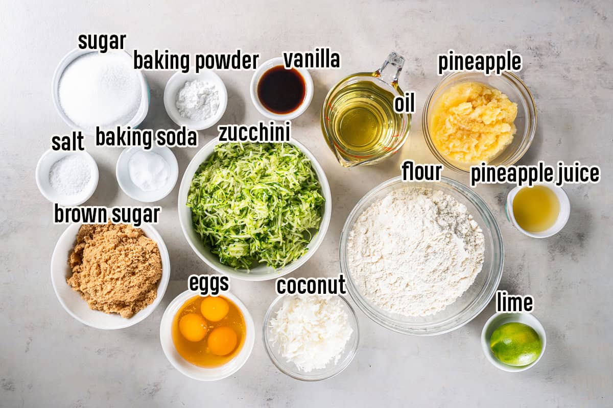 The ingredients for Pineapple Zucchini Bread arranged in bowls on a light countertop, including flour, granulated sugar, brown sugar, baking powder, baking soda, salt, eggs, vegetable oil, vanilla extract, shredded zucchini, crushed pineapple, reserved pineapple juice, sweetened shredded coconut, and a lime.
