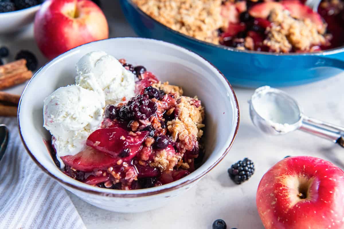 Apple Berry Crisp served warm in a bowl with two scoops of vanilla ice cream, with the baked crisp, apples, and berries in the background.
