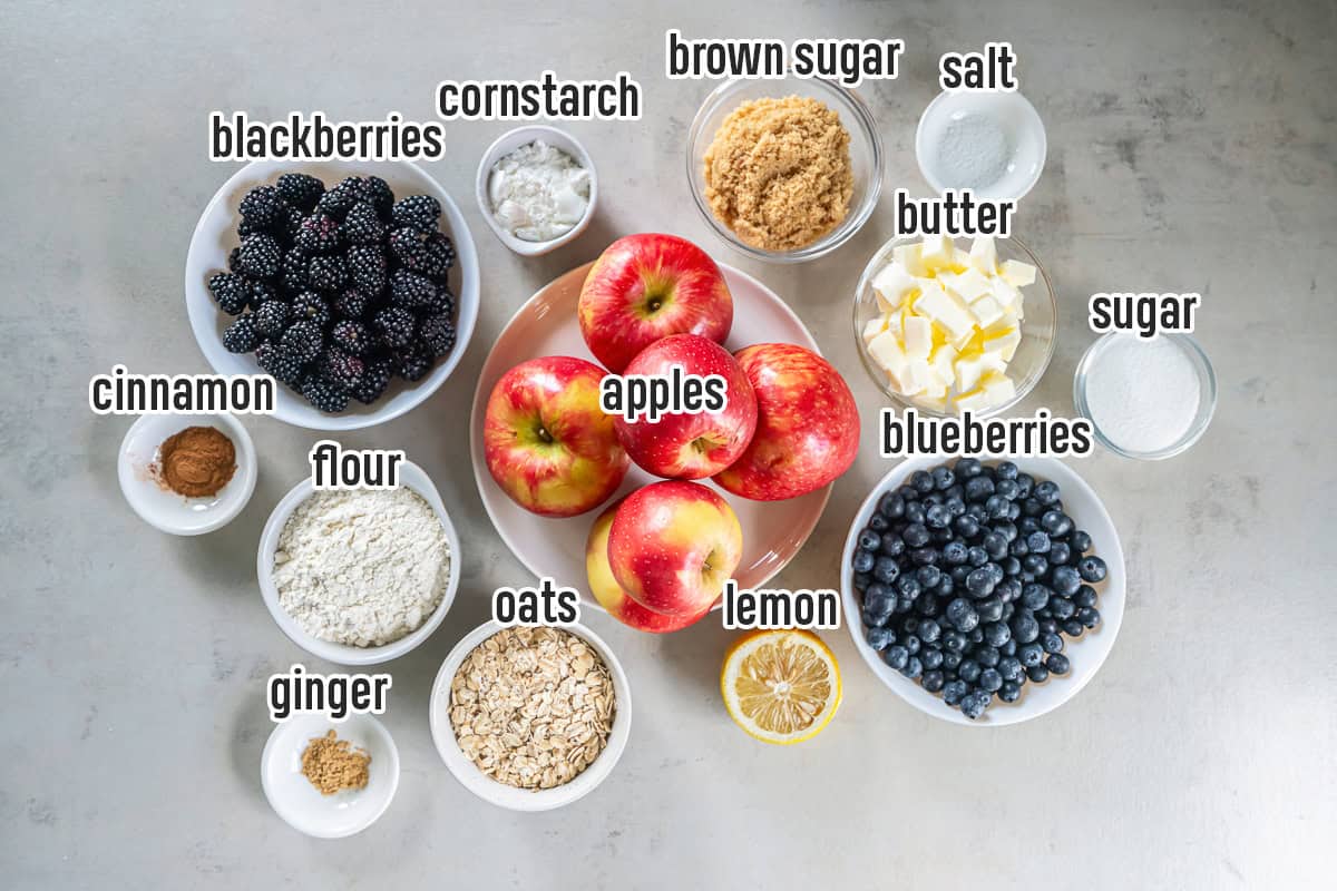 Ingredients for Apple Berry Crisp arranged on a countertop, including apples, blueberries, blackberries, lemon, oats, flour, brown sugar, granulated sugar, butter, cornstarch, cinnamon, ginger, and salt.