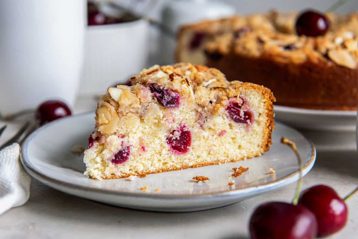 Slice of cherry coffee cake with fresh cherries and almond streusel topping on a plate, with the whole cake in the background.