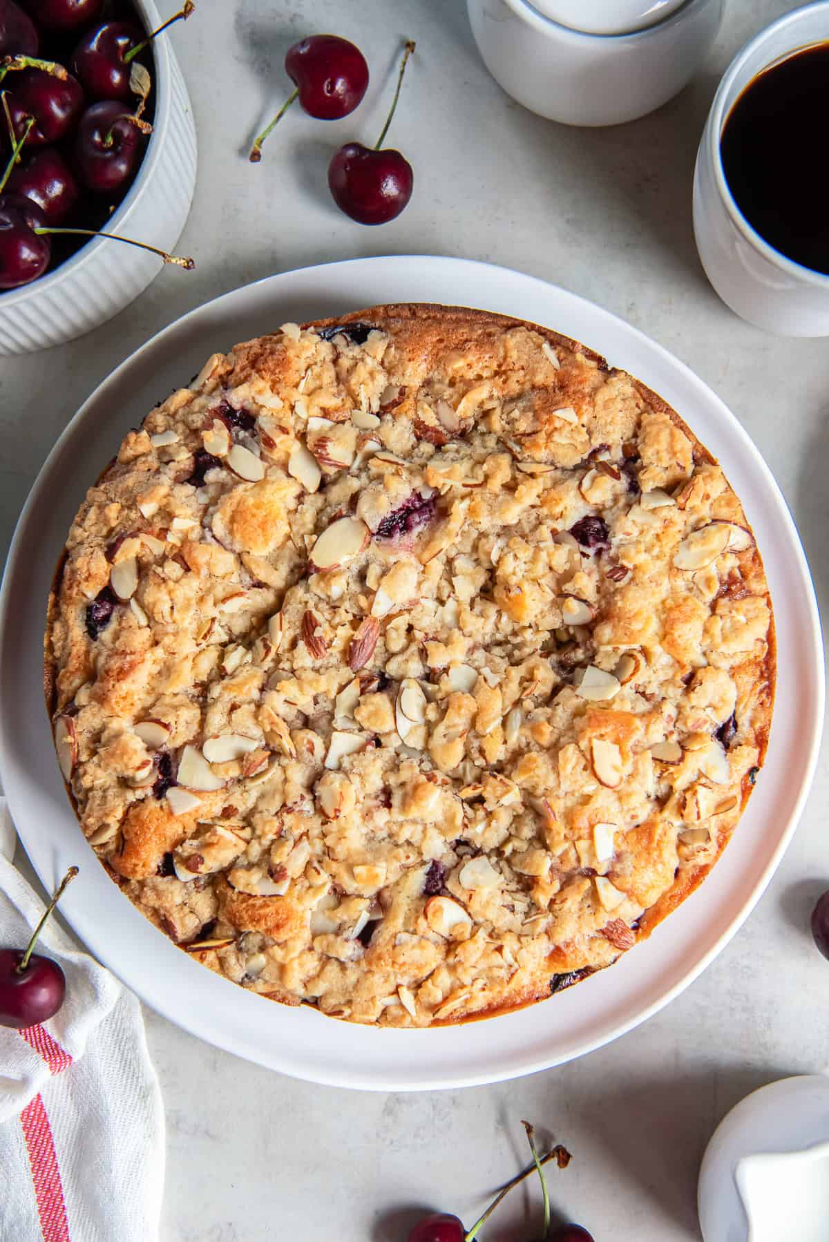 A round cherry coffee cake topped with buttery almond streusel on a white serving platter, surrounded by fresh cherries and a cup of coffee.