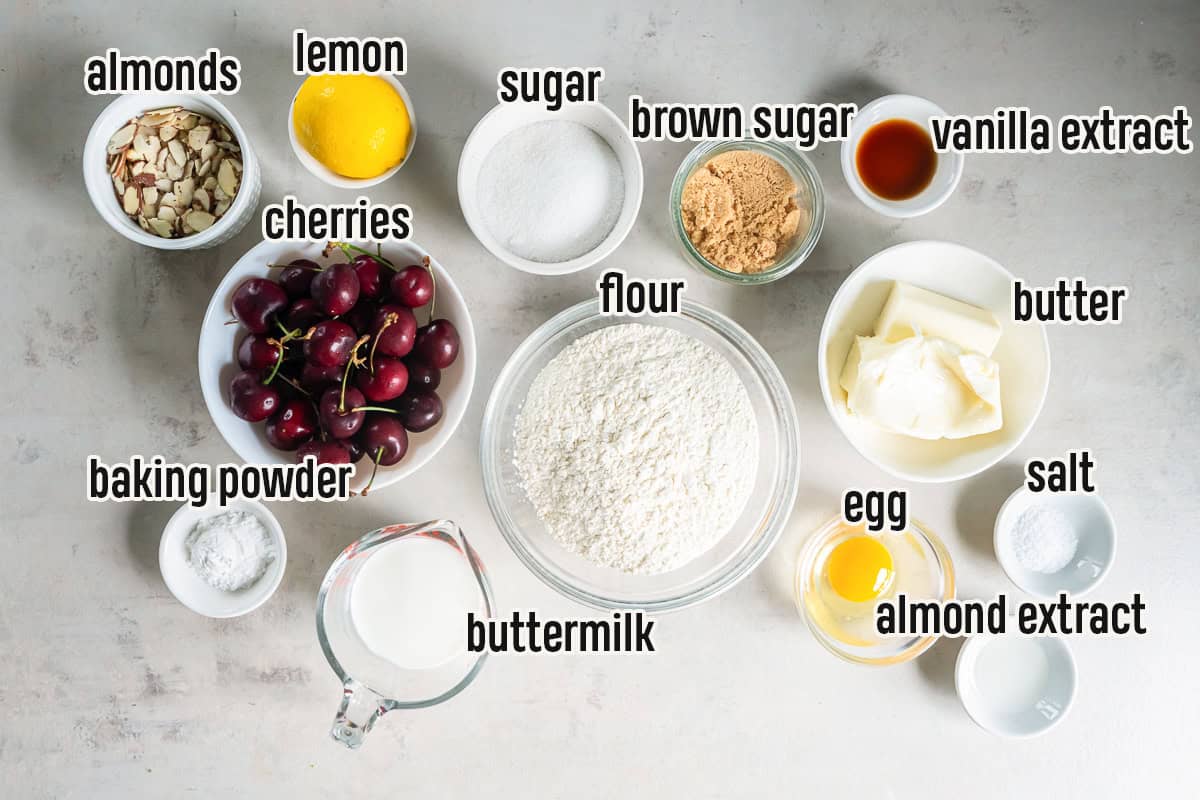 Ingredients to make cherry coffee cake arranged on a countertop, including fresh cherries, flour, butter, buttermilk, sliced almonds, lemon, sugars, egg, extracts, baking powder, and salt.