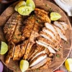 Grilled chili lime chicken breasts, some sliced, on a wooden serving board with lime wedges, tortillas, corn, guacamole, and tomatoes.