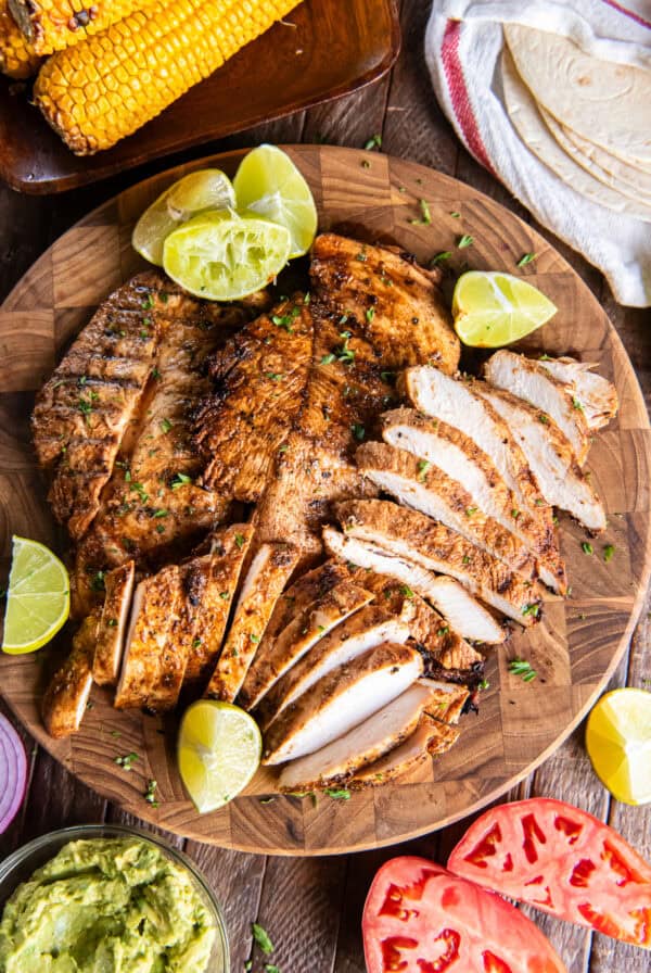 Grilled chili lime chicken breasts, some sliced, on a wooden serving board with lime wedges, tortillas, corn, guacamole, and tomatoes.
