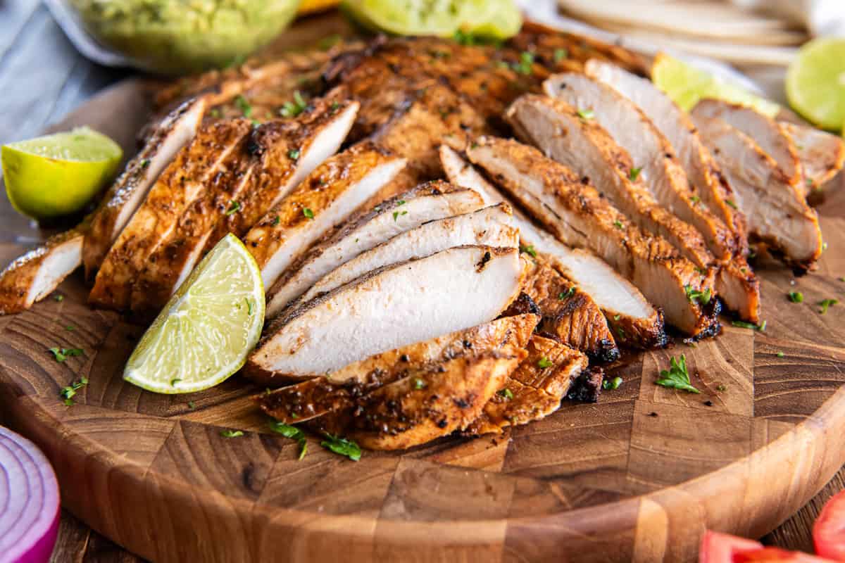 Sliced chili lime chicken breasts on a wooden serving board with fresh lime wedges.