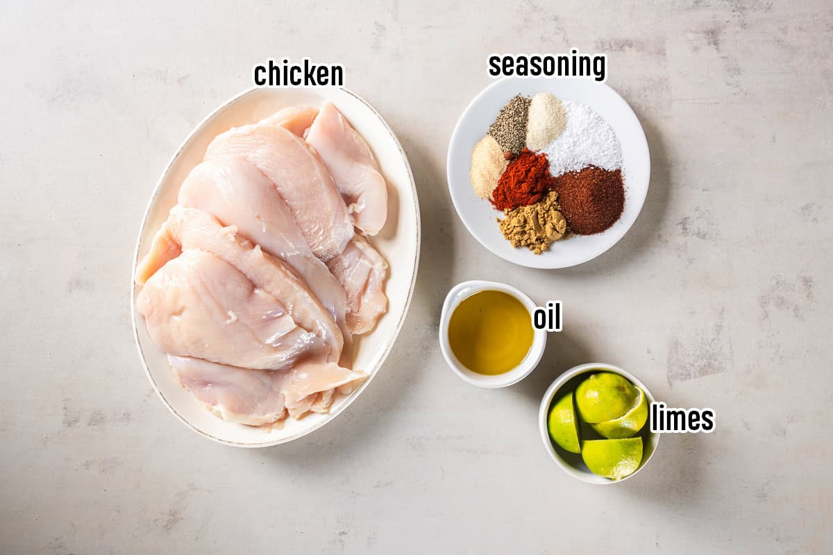 Ingredients for chili lime chicken including thin-sliced chicken breasts, olive oil, lime wedges, and a plate of spices labeled chicken, oil, limes, and seasoning.