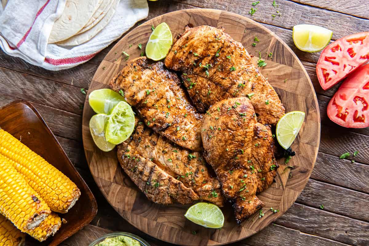 Grilled chili lime chicken breasts with browned edges and grill marks on a sheet pan surrounded by corn on the cob and tomato wedges.