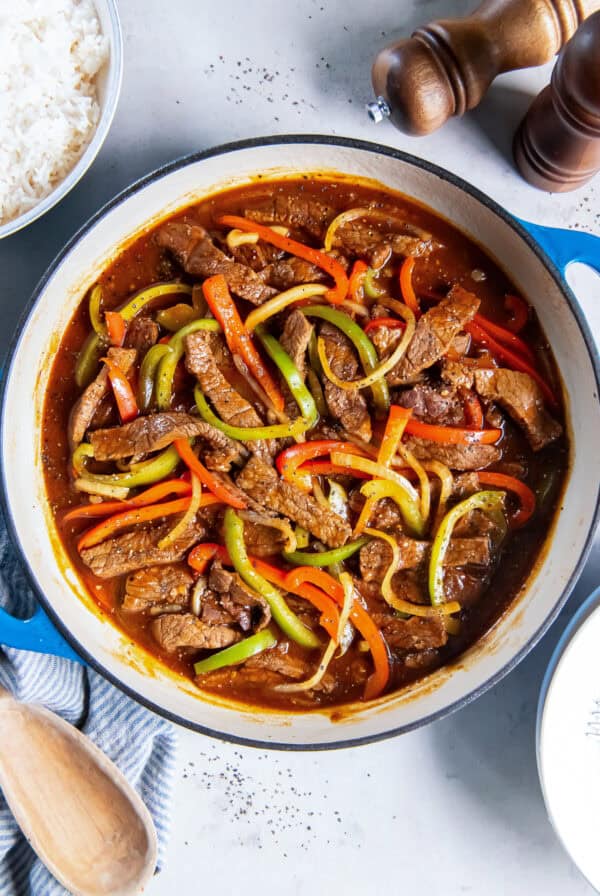 Easy Pepper Steak made with tender beef, bell peppers, and onion simmered in a flavorful brown sauce, served in a blue skillet with white rice on the side.