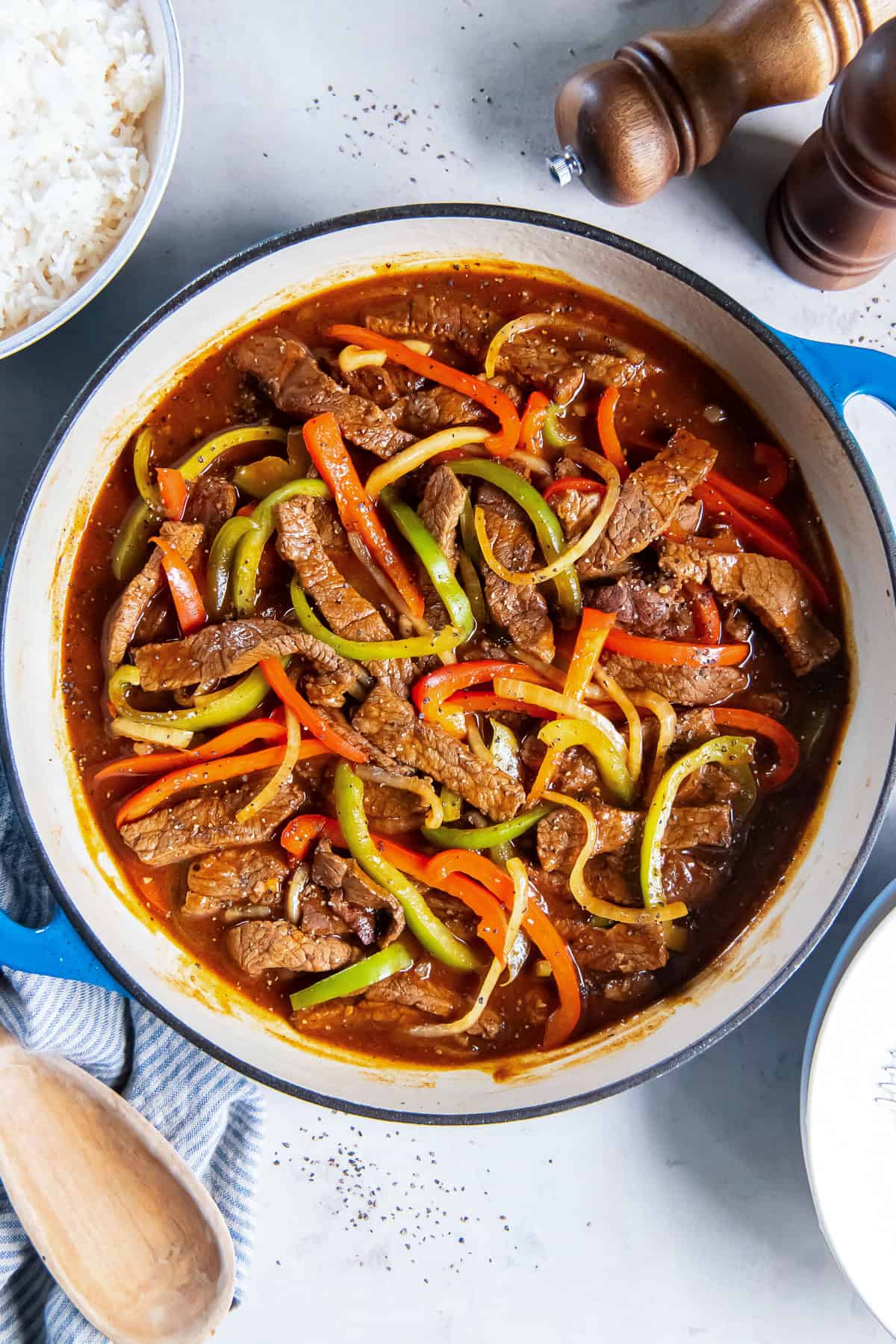 Easy Pepper Steak made with tender beef, bell peppers, and onion simmered in a flavorful brown sauce, served in a blue skillet with white rice on the side.