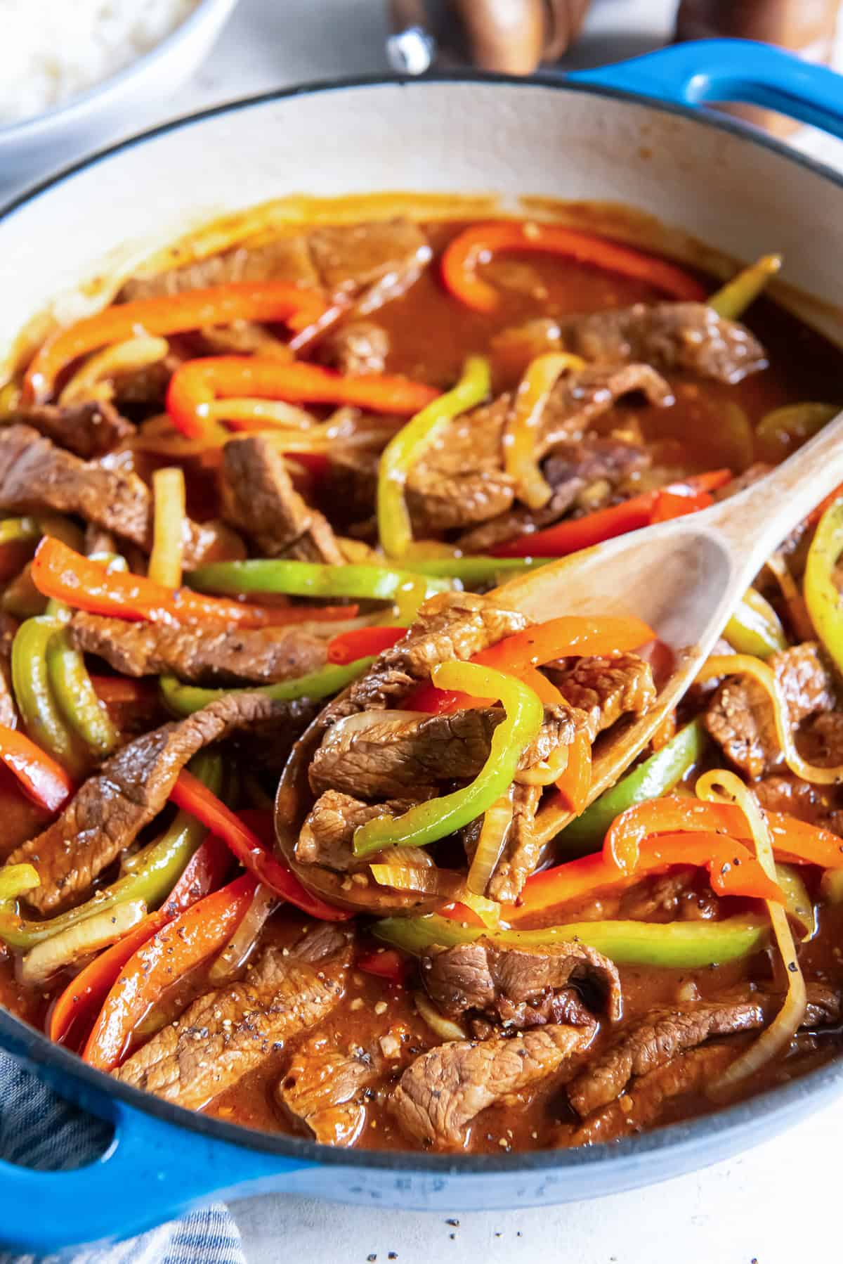 Wooden spoon lifting Pepper Steak with tender strips of beef, colorful bell peppers, and onion from a skillet filled with rich brown sauce.