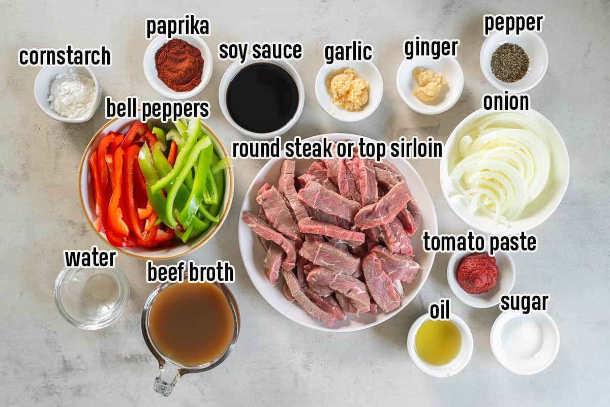 Ingredients for Pepper Steak arranged on a countertop, including sliced round steak, bell peppers, onion, beef broth, soy sauce, paprika, tomato paste, garlic, ginger, cornstarch, oil, sugar, black pepper, and water.