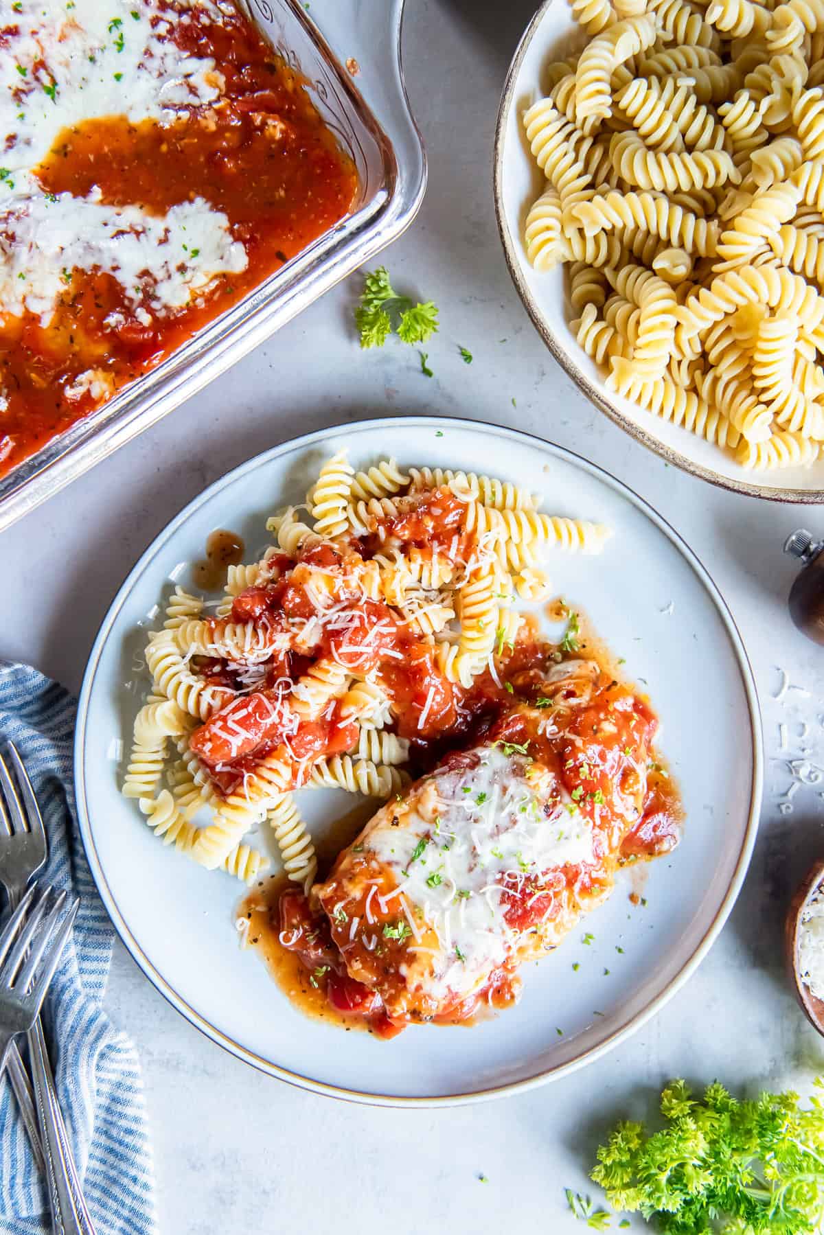 Plate of Easy Italian Chicken topped with melted mozzarella and tomato sauce, served with rotini pasta and grated Parmesan, with the baking dish in the background.